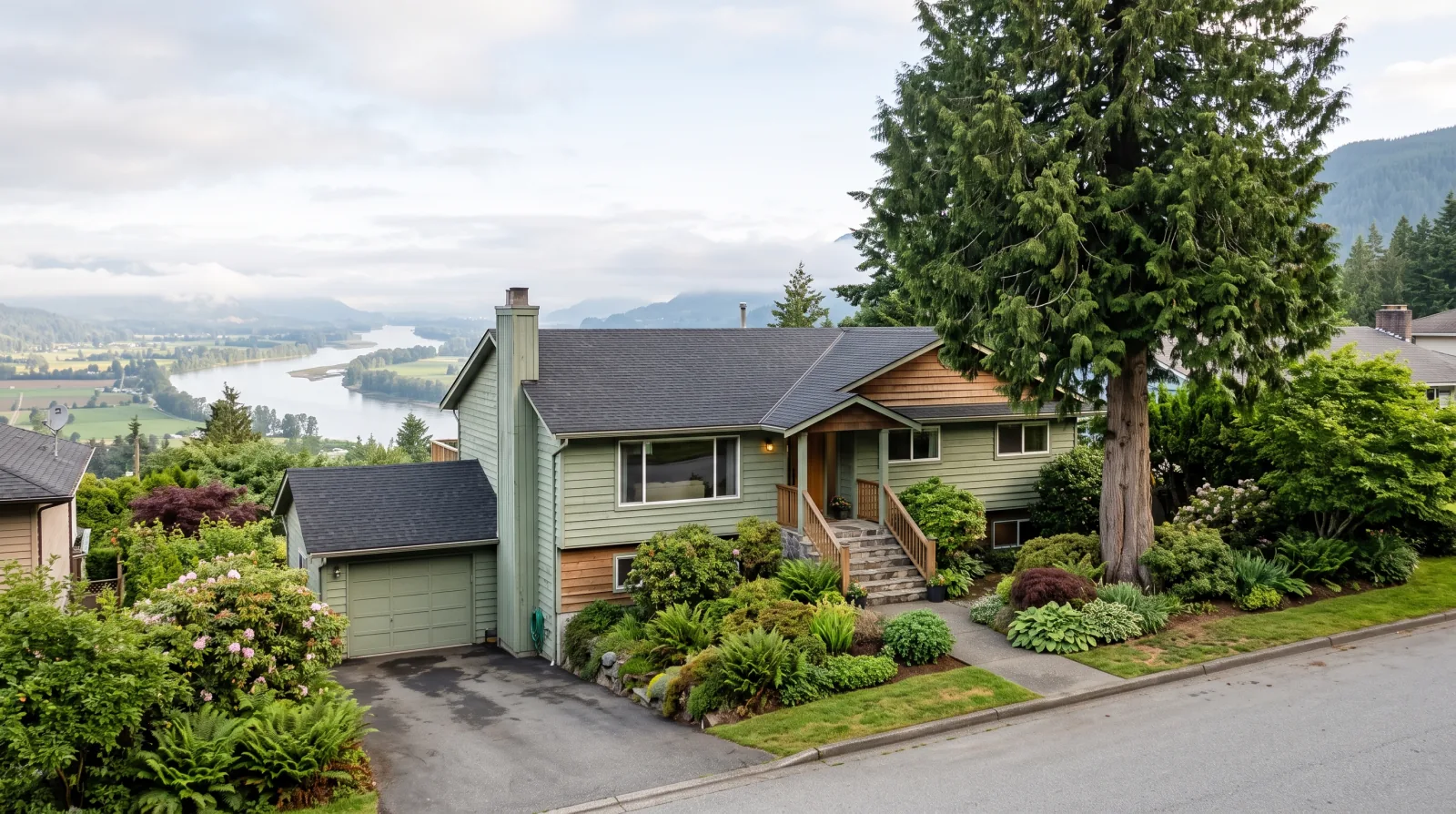 A split-level house with green siding, an attached garage and a tall evergreen tree on a hillside street, with a wide river and farmland in the valley below